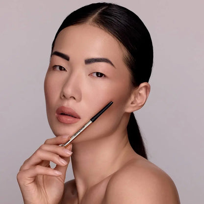 Anwendung MoniAmori Extra-LipStay MM033 Blush Sand: Model hält den roségoldenen Automatikstift seitlich ans Gesicht, Nahaufnahme mit sichtbarem Lipliner in Blush Sand, grauer Hintergrund.