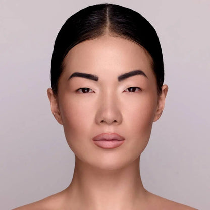 Model mit MoniAmori Extra-LipStay MM033 Blush Sand: Frau mit dunklem zurückgekämmtem Haar und braunen Augen, natürlichem Make-up und Lippenstift in zartem Blush Sand, frontale Porträtaufnahme vor grauem Hintergrund.