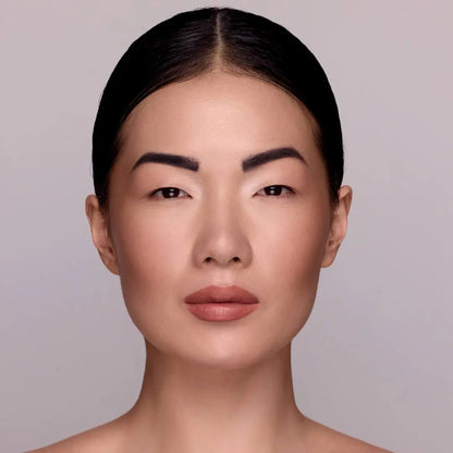 Model mit MoniAmori Extra-LipStay MM033 Blush Sand: Frau mit dunklem zurückgekämmtem Haar, intensiverem Lippenmakeup in sattem Blush Sand, natürliches Porträt vor grauem Hintergrund.