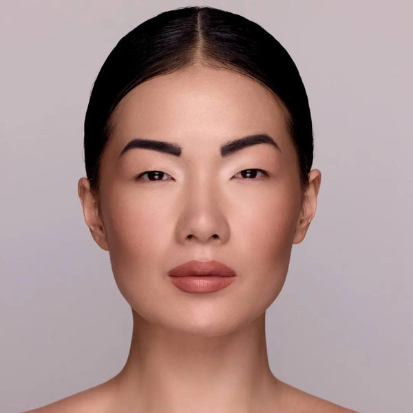 Model mit MoniAmori Extra-LipStay MM033 Blush Sand: Frau mit dunklem zurückgekämmtem Haar, intensiverem Lippenmakeup in sattem Blush Sand, natürliches Porträt vor grauem Hintergrund.